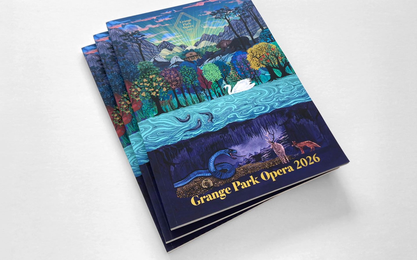 Three stacked Grange Park Opera 2026 programme booklets showing the Das Rheingold cover illustration by Shadric Toop, depicting a mountain landscape with a glowing opera house, forests, river, and symbolic animals.